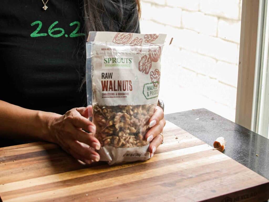 A person wearing a black shirt holds a bag of Sprouts Farmers Market raw walnuts on a wooden cutting board, with some walnut pieces scattered nearby.